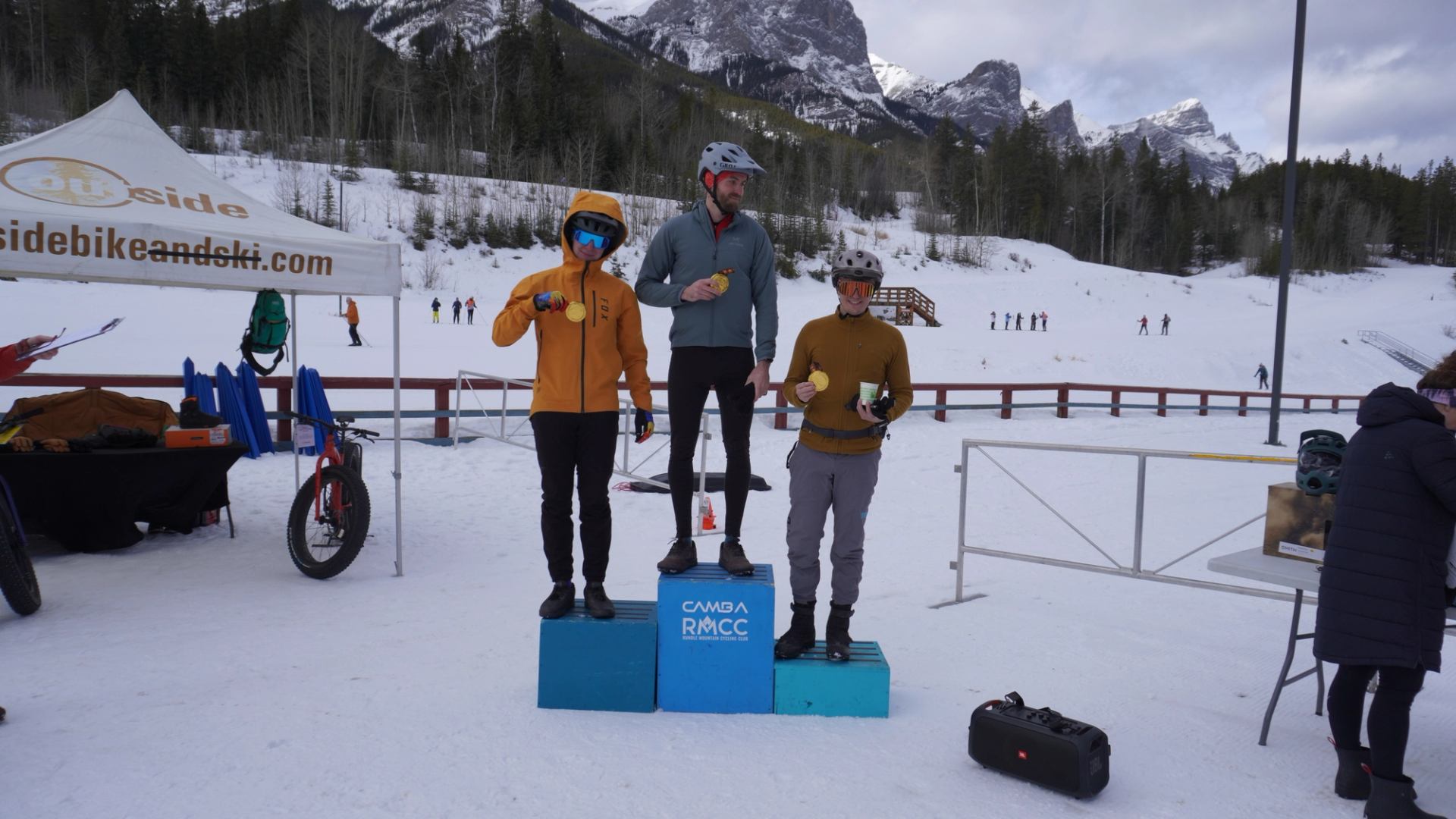 Three riders standing on a snowy podium holding awards with mountains and event tents behind them.