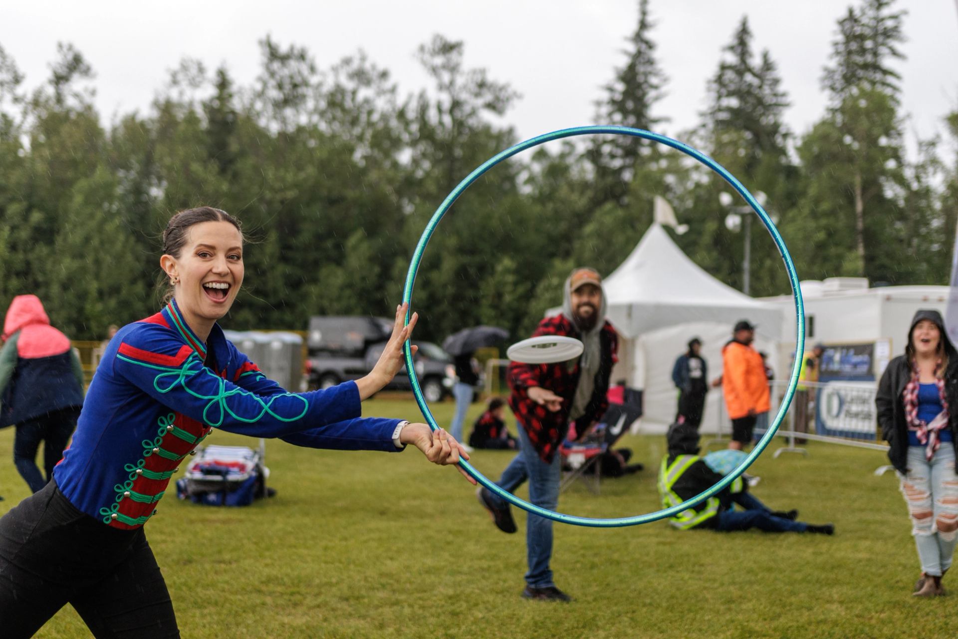 Performer holding a hoop while someone tosses a disc through it at an outdoor festival.