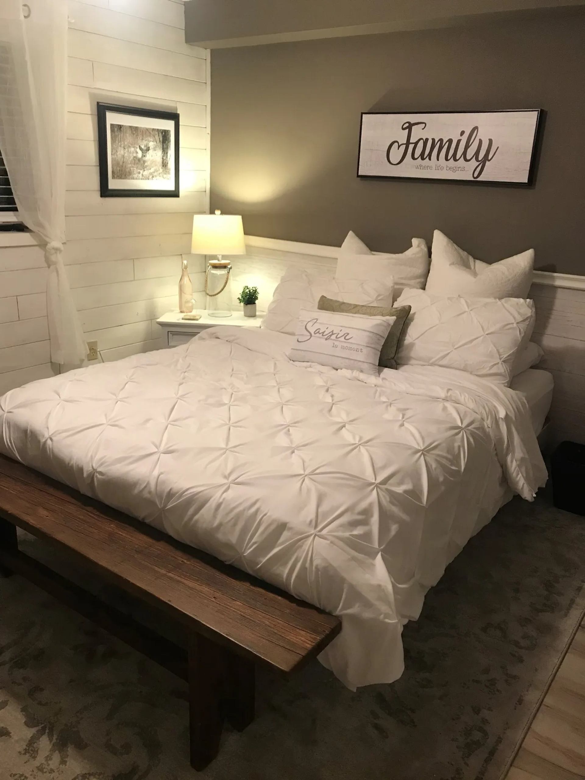 Bedroom with white bedding, wooden bench, bedside lamp and framed wall signs.