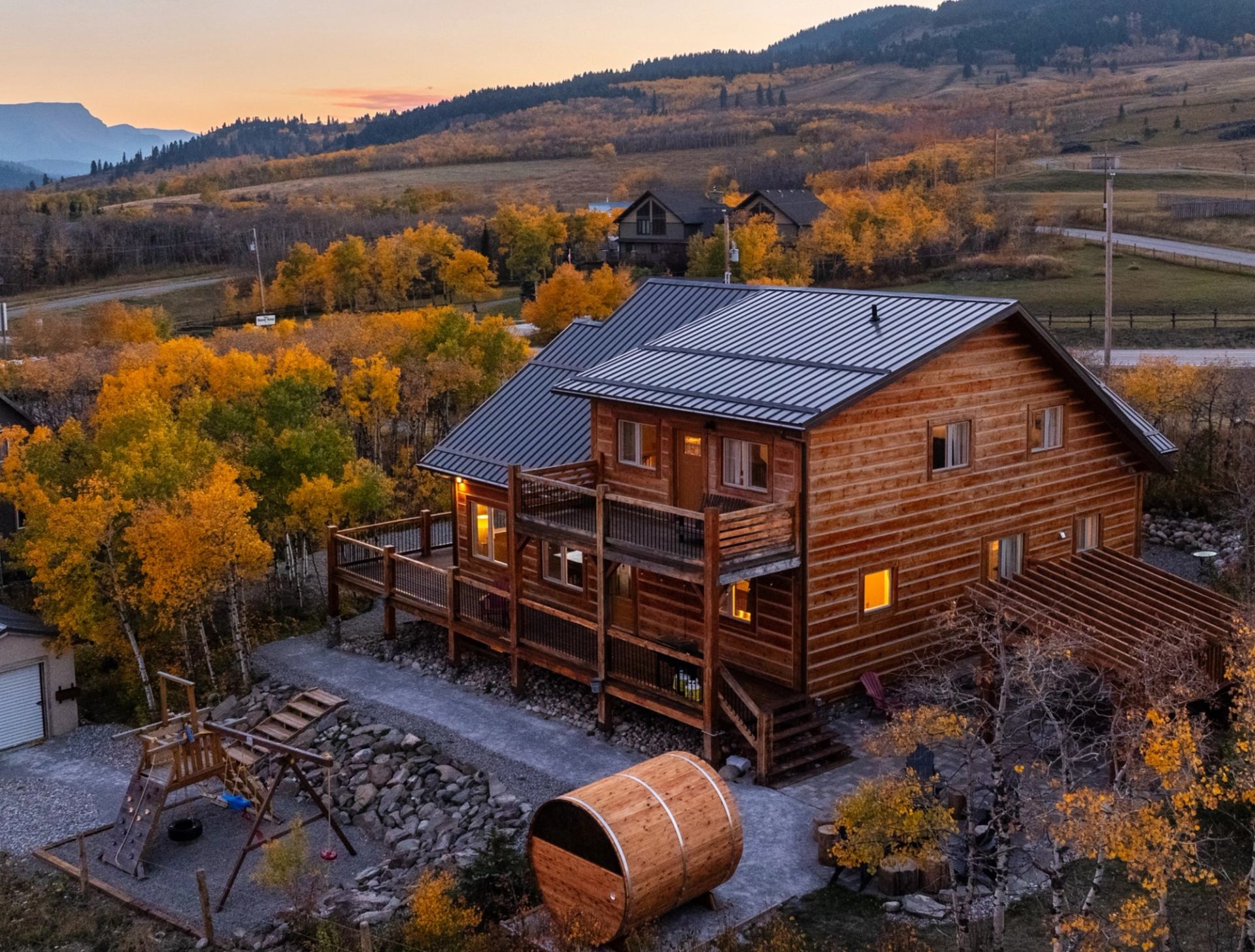 Large wooden cabin with deck, surrounded by fall trees and mountains at sunset.