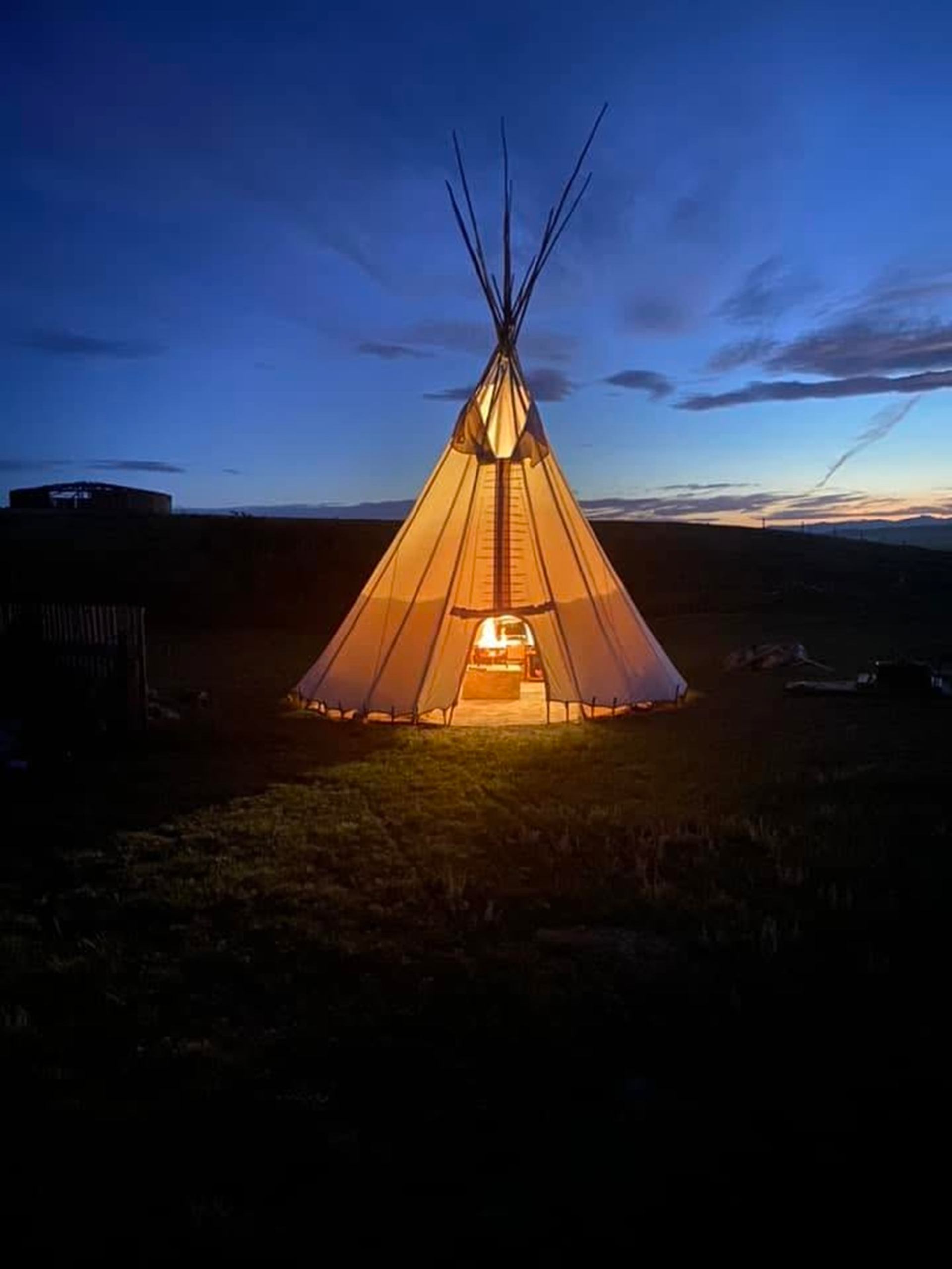 Illuminated tipi on open grassland under deep blue evening sky with scattered clouds.