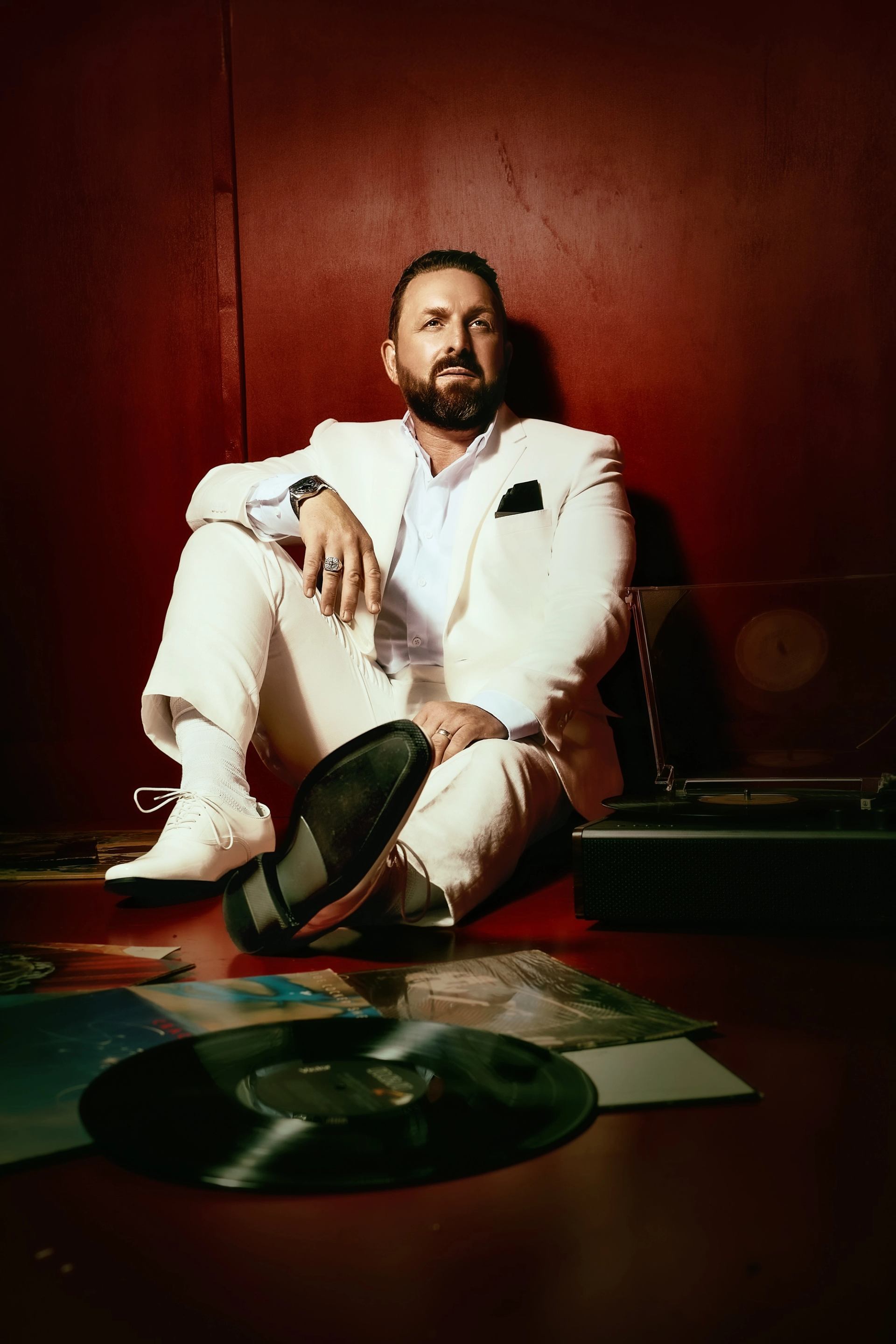 Bearded person in a white suit sits beside a record player and scattered vinyl records.