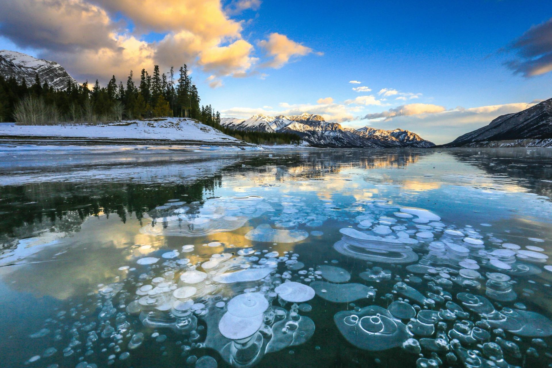 Frozen lake with trapped bubbles reflects clouds beneath snow-covered mountains.