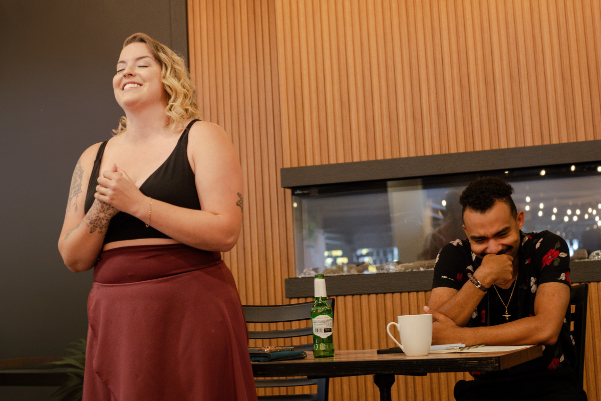 Person standing and speaking beside seated person at table in modern wood-panelled room.