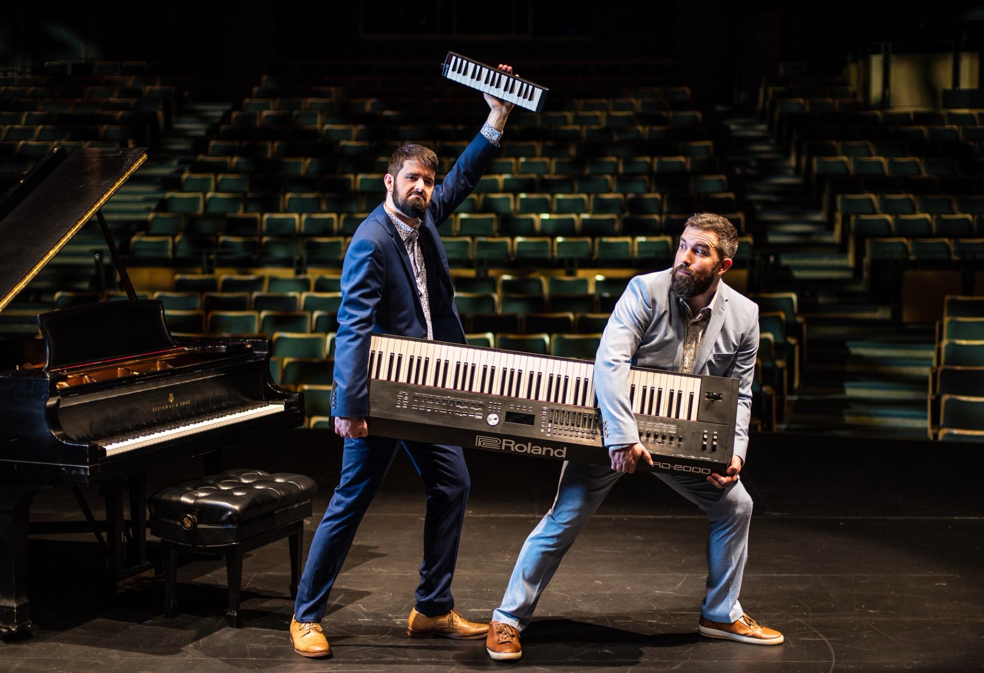 Two suited performers pose with large Roland keyboards on a theatre stage beside grand piano.