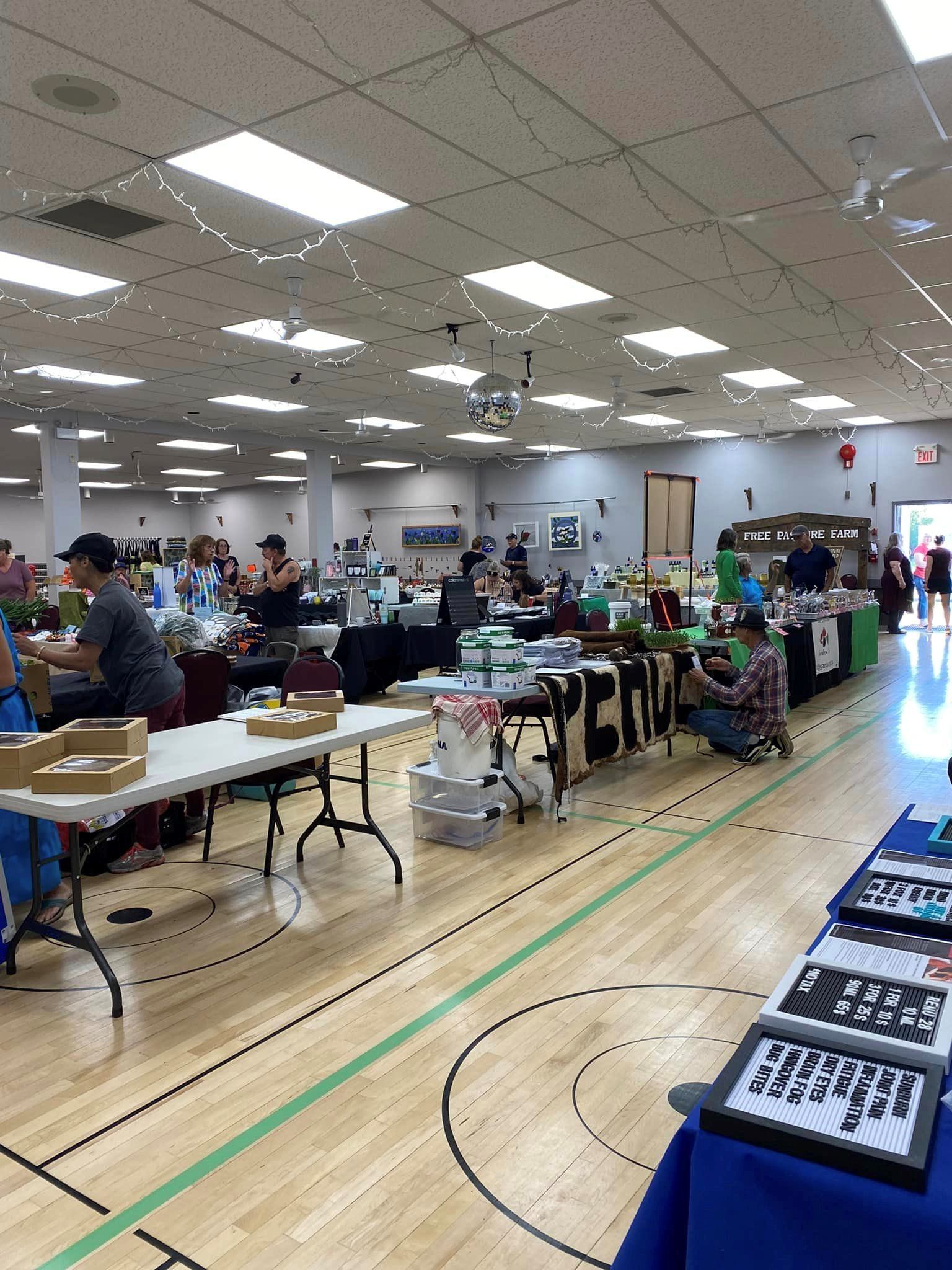 Indoor farmers’ market with multiple vendor tables and shoppers in a bright hall.