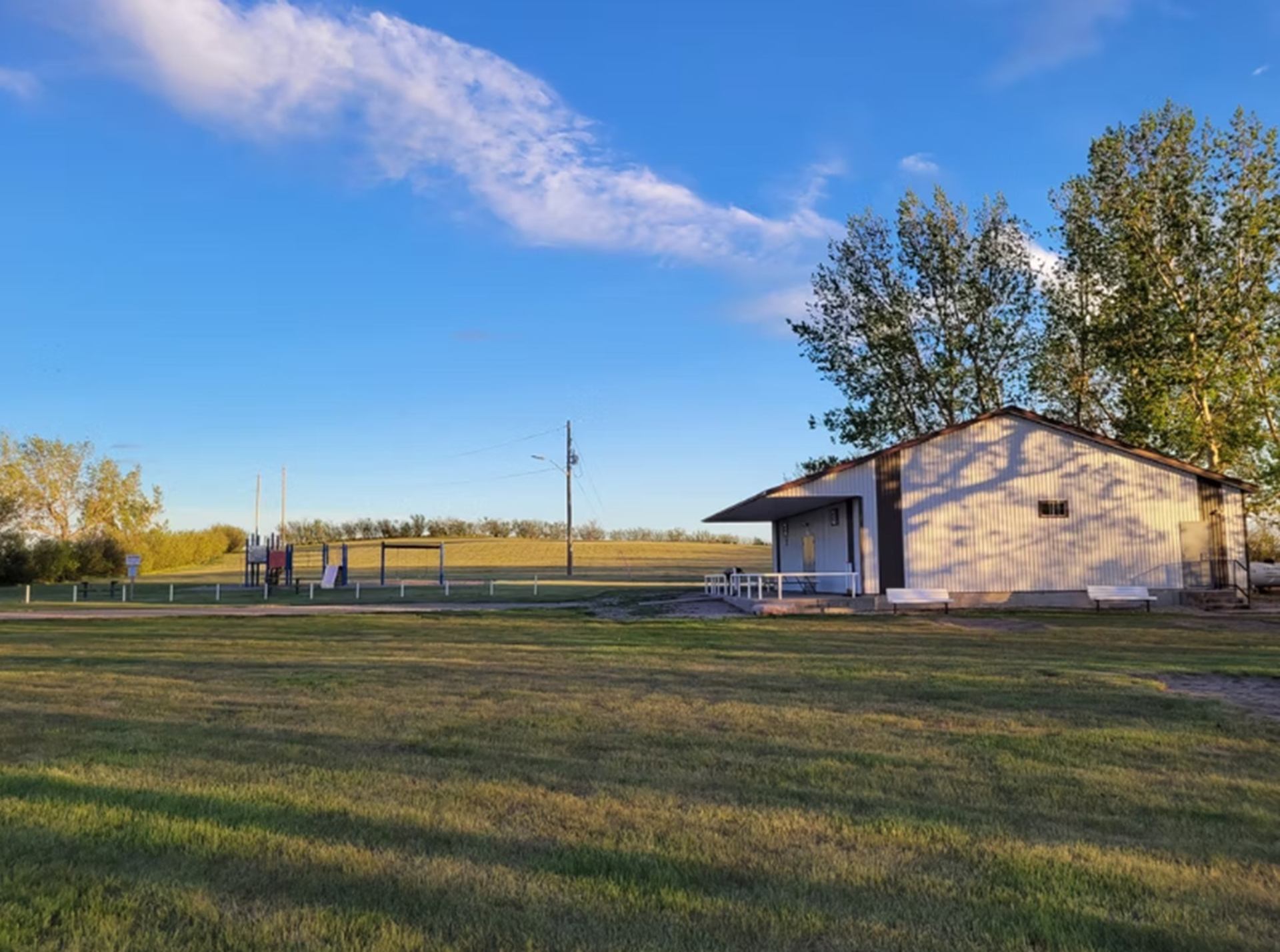 Small park building beside open grassy field, playground and trees under blue sky.