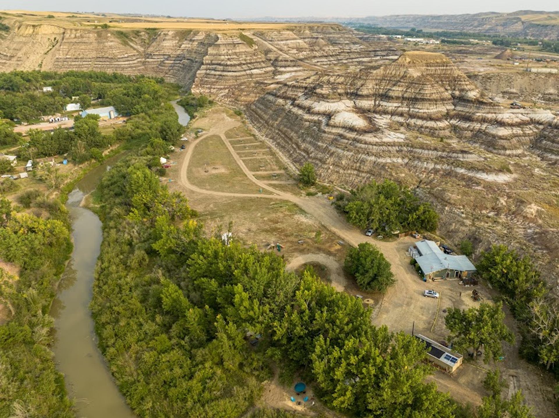Aerial view of river, campground and buildings beside layered badlands cliffs and trees.