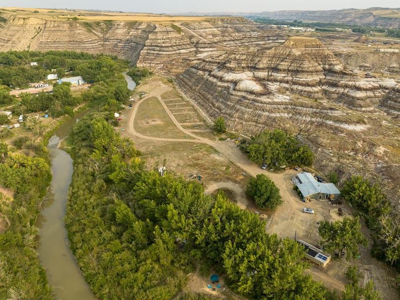Aerial view of river, campground and buildings beside layered badlands cliffs and trees.