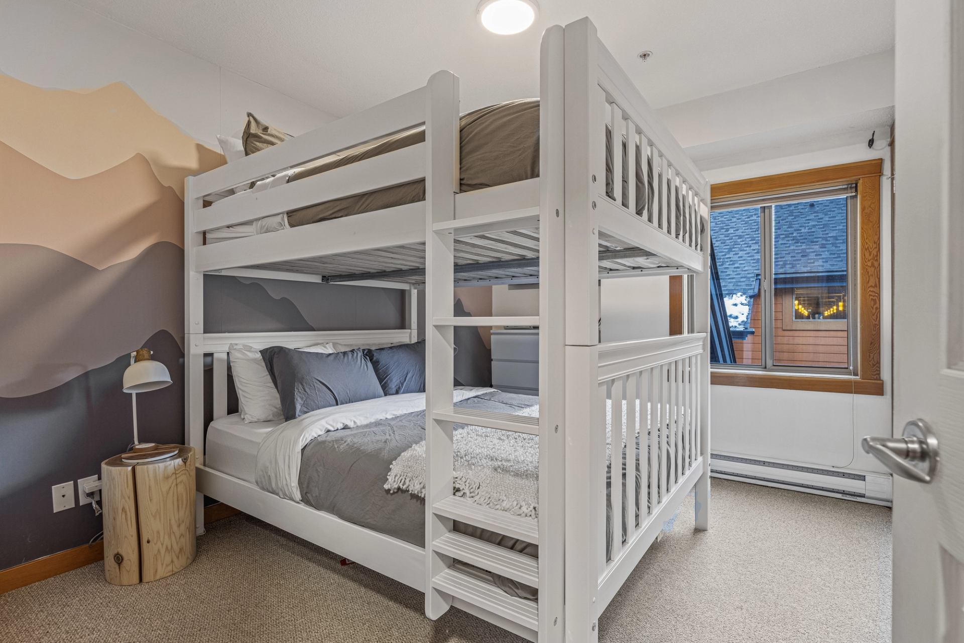 Bedroom with white bunk bed, mountain mural wall, small log nightstand and window.