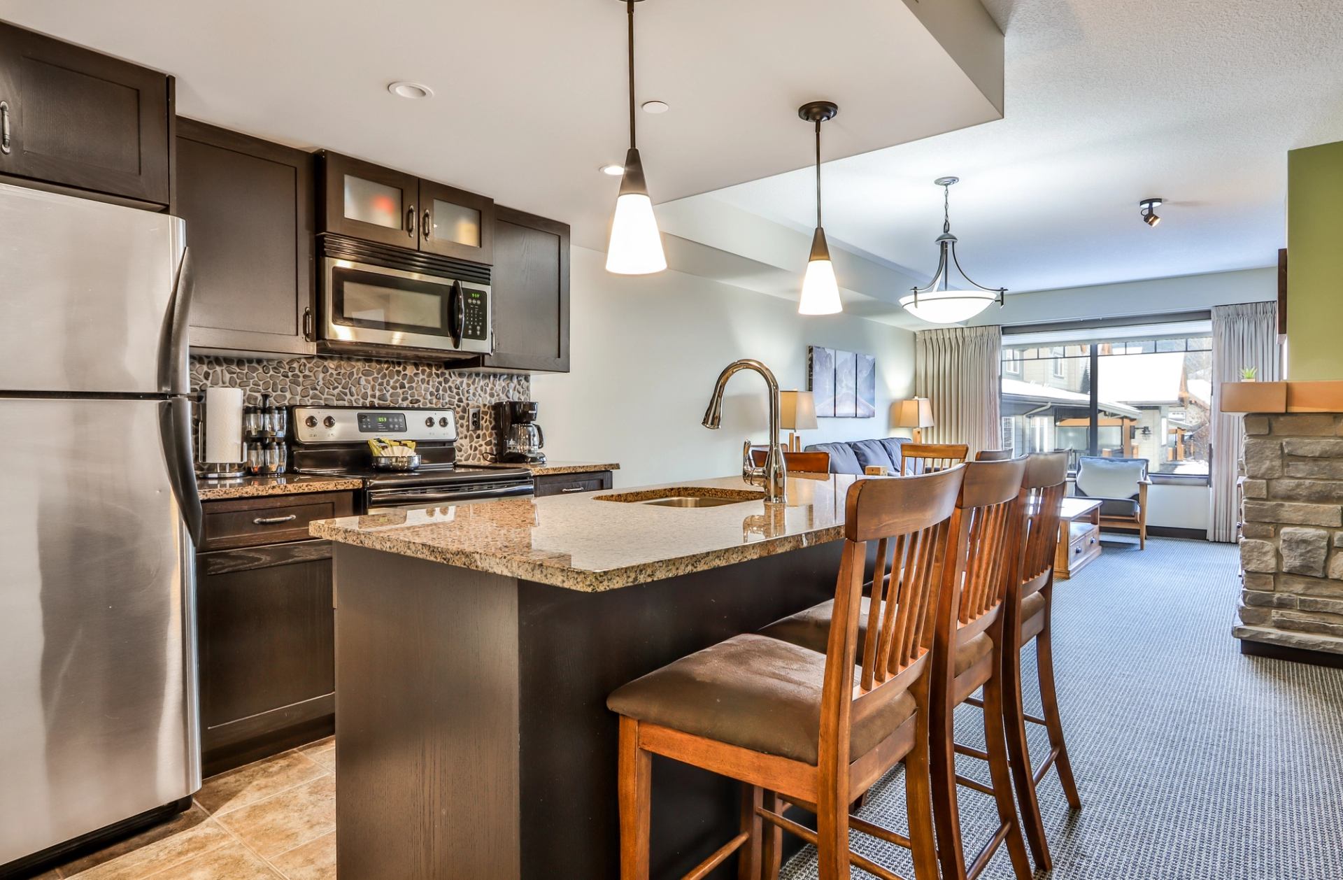 Modern open-concept kitchen and living room with dark cabinets, stainless steel appliances and a stone fireplace.