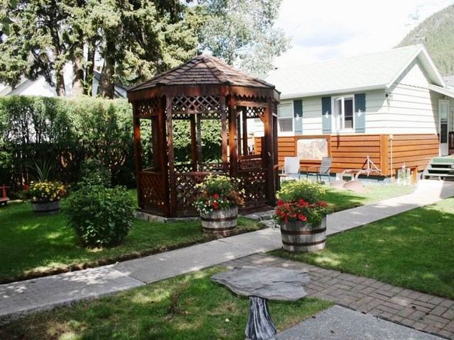 A tranquil garden scene with a wooden gazebo surrounded by lush greenery and colorful flower pots. A cozy house sits in the background, under a blue sky.