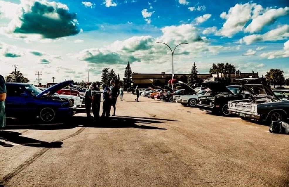 Classic cars with open hoods line a parking lot as people talk and look.