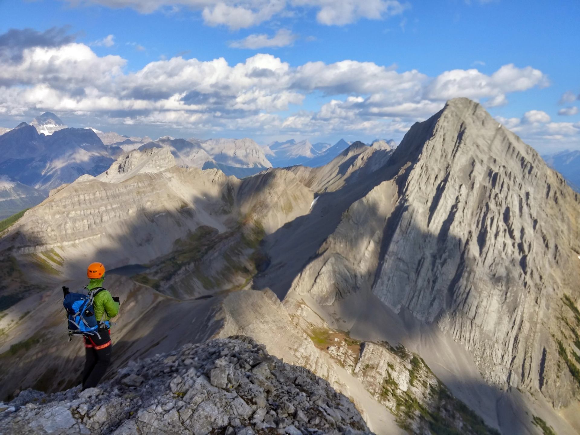 Hiker in orange helmet stands on rocky ridge overlooking dramatic grey mountain peaks.