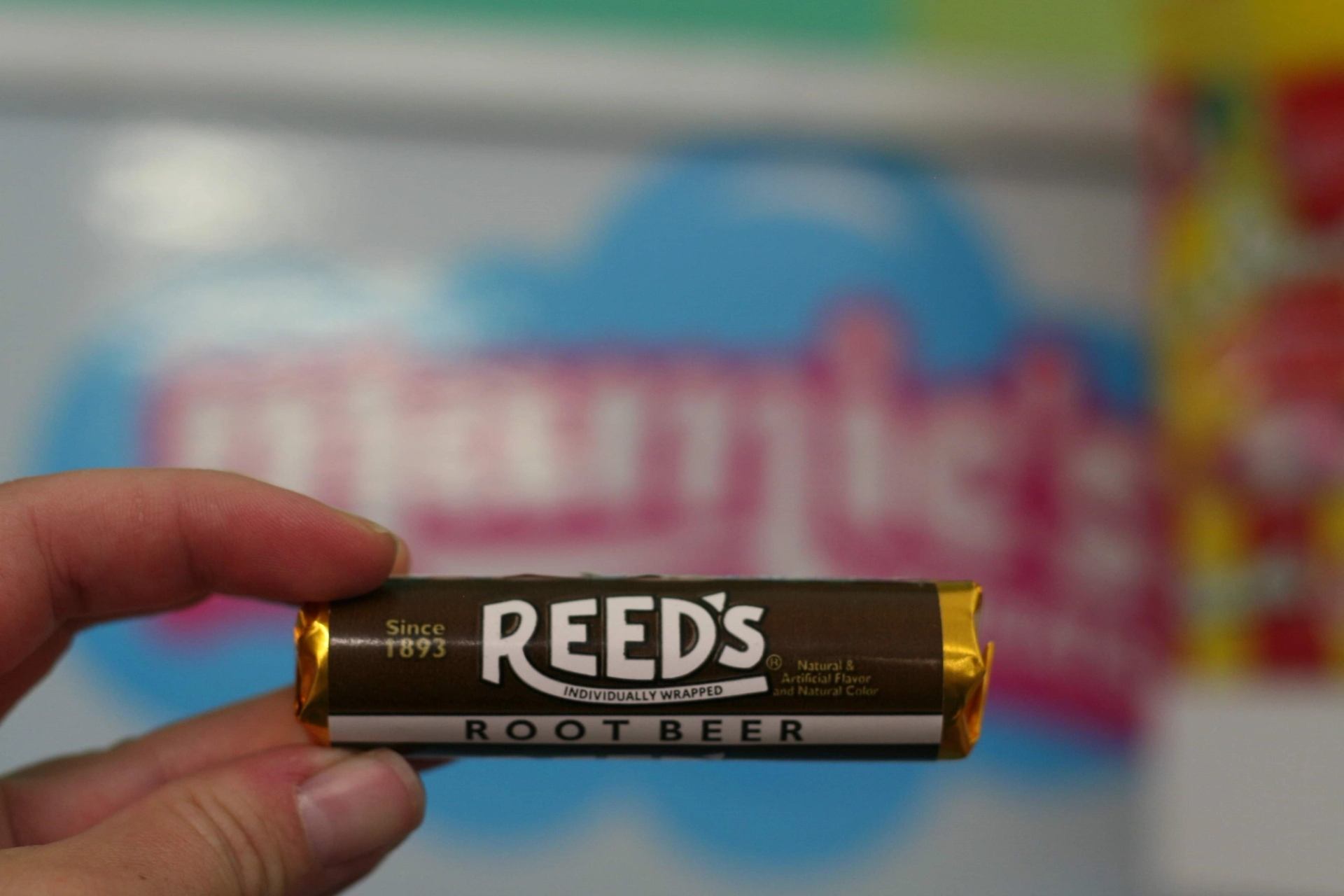 Hand holding a Reed’s Root Beer candy roll in front of a colorful shop display.