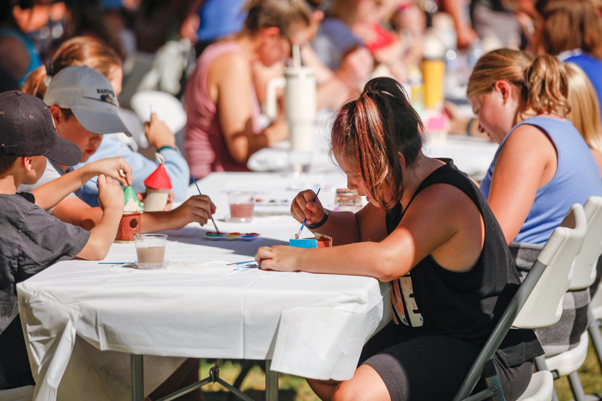 Children and adults sitting at tables outdoors, painting small crafts during a community activity.