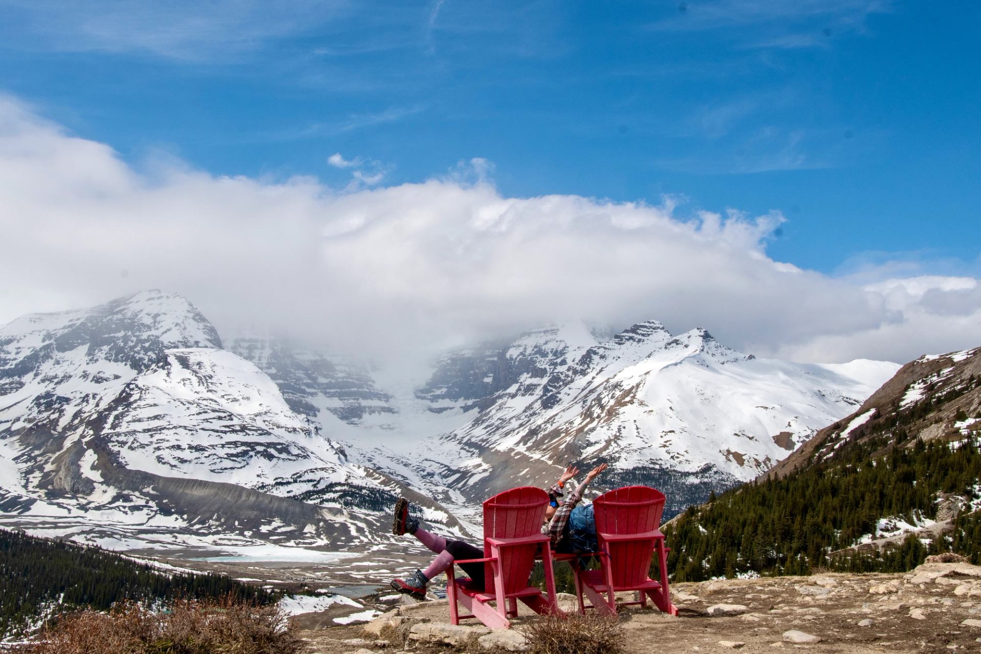Two people lounging in red chairs overlooking snowy mountain peaks and a wide valley.