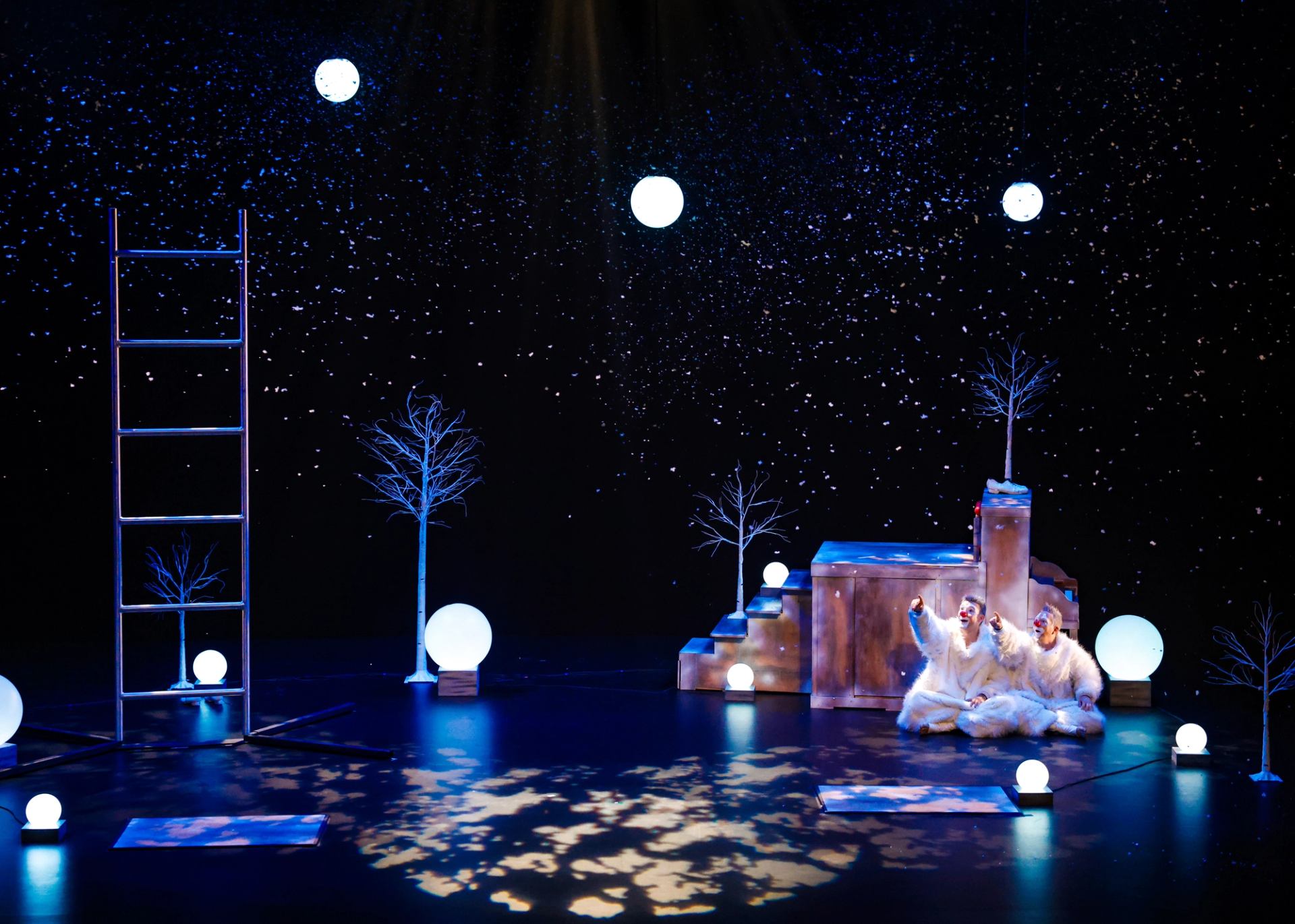 Theatre stage with snowy set, glowing orbs, bare trees and two performers in white costumes.