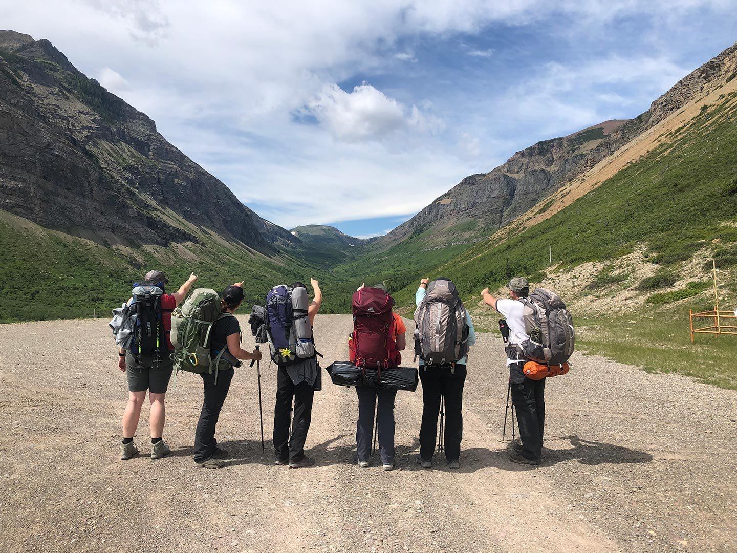 Backpackers stand on gravel road between steep green mountains, pointing toward distant valley.
