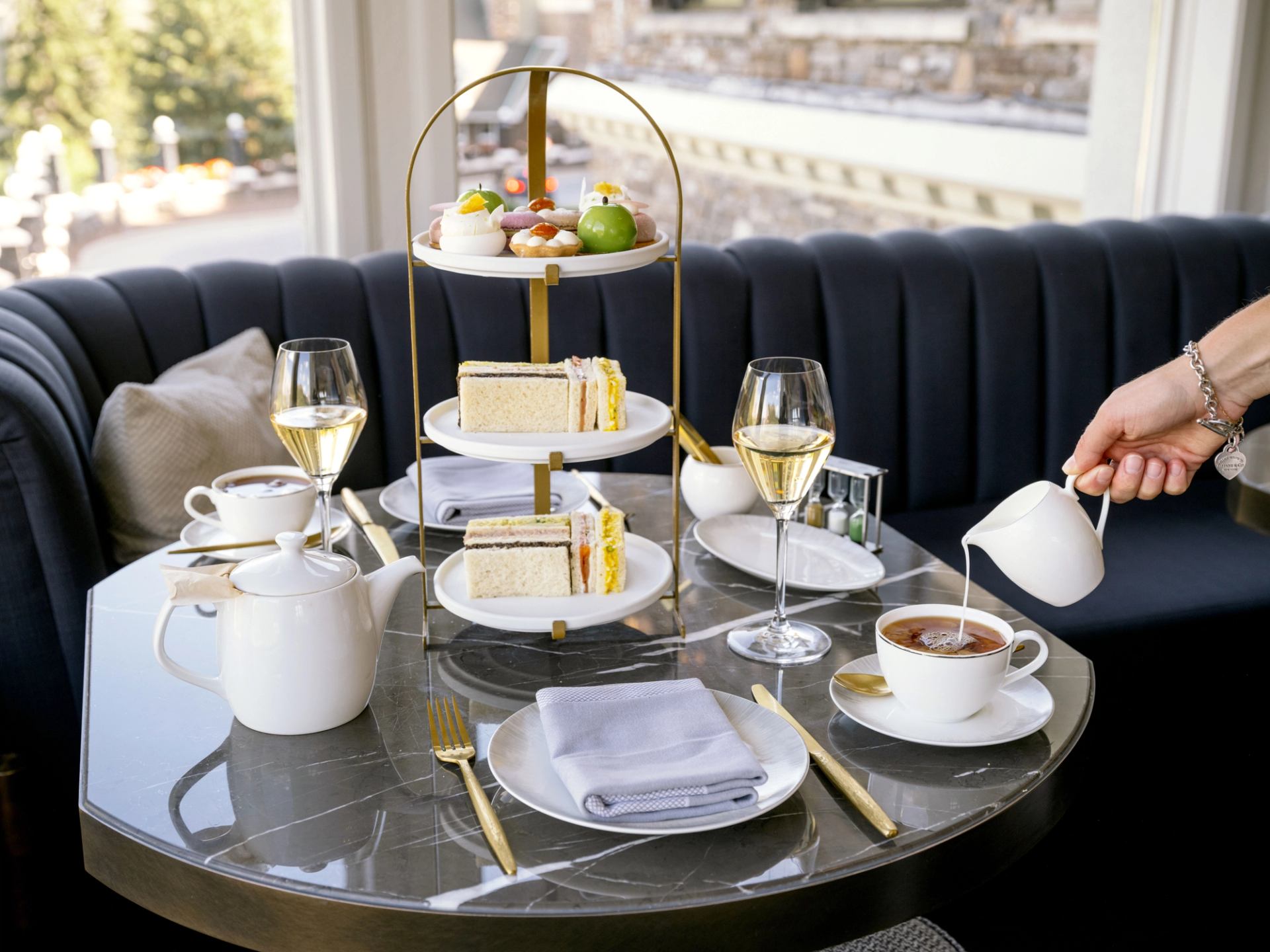 A hand pours milk into tea at an elegant table set for afternoon tea, with a tiered stand of sandwiches and pastries.