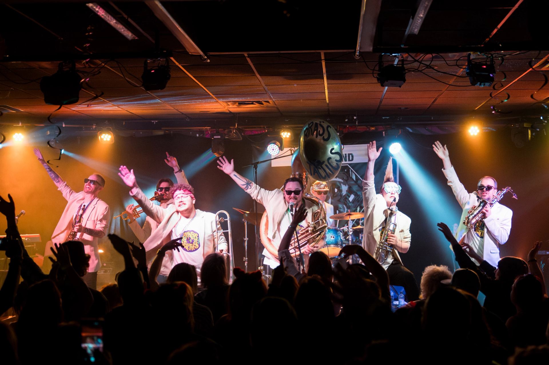 Brass band performs on stage with raised arms as crowd silhouettes cheer below.