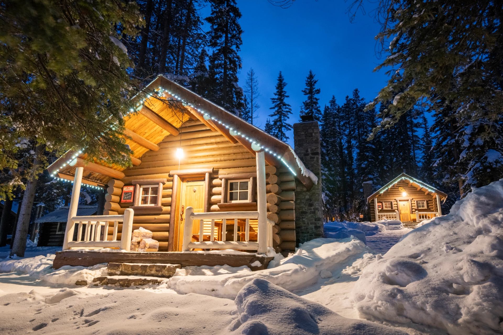 Two lit log cabins in a snowy forest at dusk.