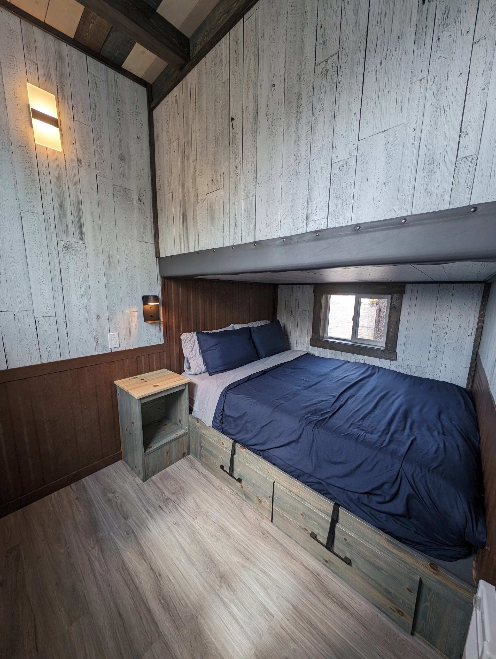 Compact bedroom with blue bedding, wood-panelled walls, wall lights and a small window.