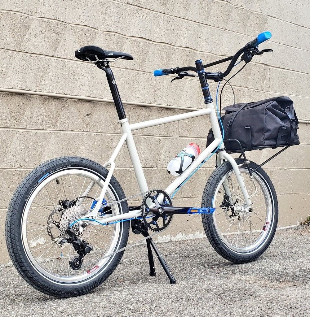 White bike with blue accents and rear bag parked by a beige brick wall.