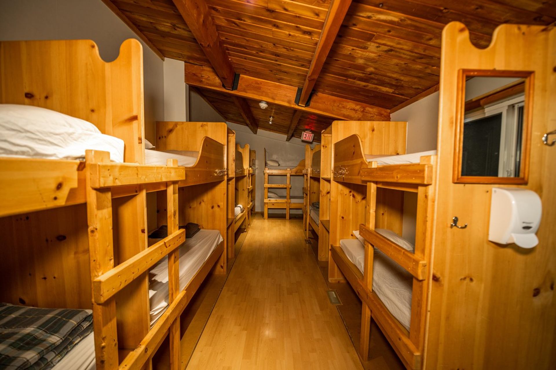Shared dorm room with multiple wooden bunk beds lining both sides, vaulted ceiling.