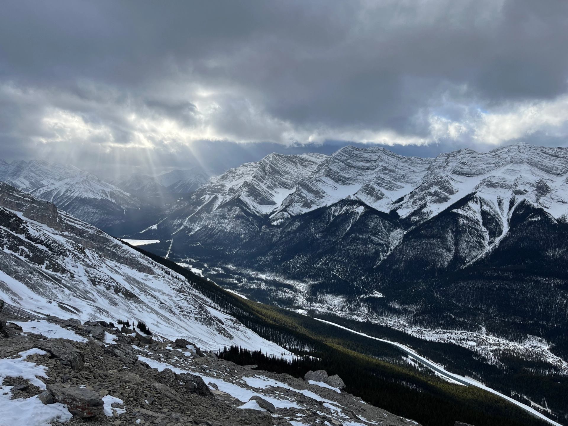 Snow-covered mountain range with rocky slope, forested valley and sunbeams breaking through clouds.