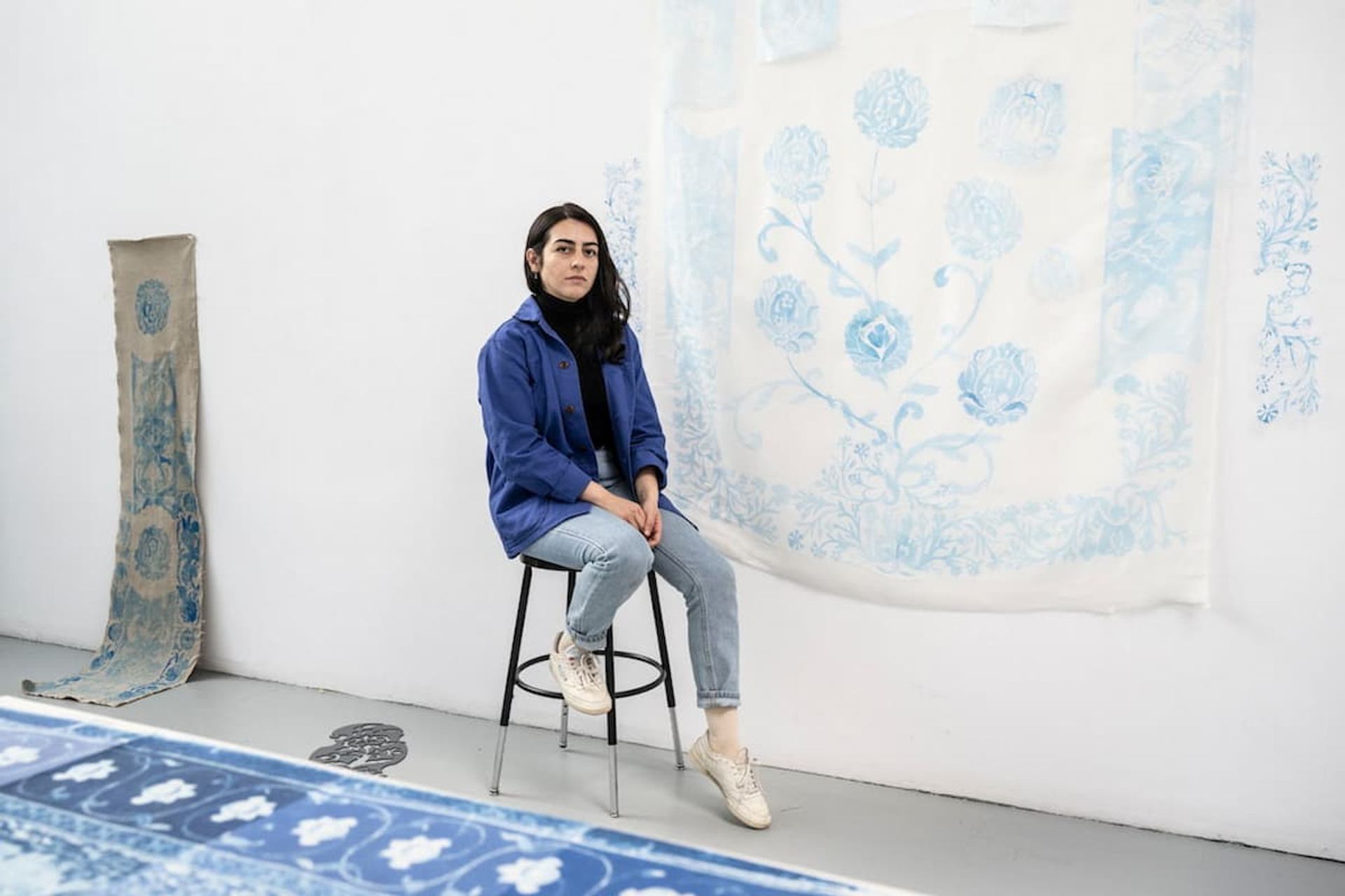 An artist sits on a stool in a white studio beside blue patterned textile works displayed on the wall and floor