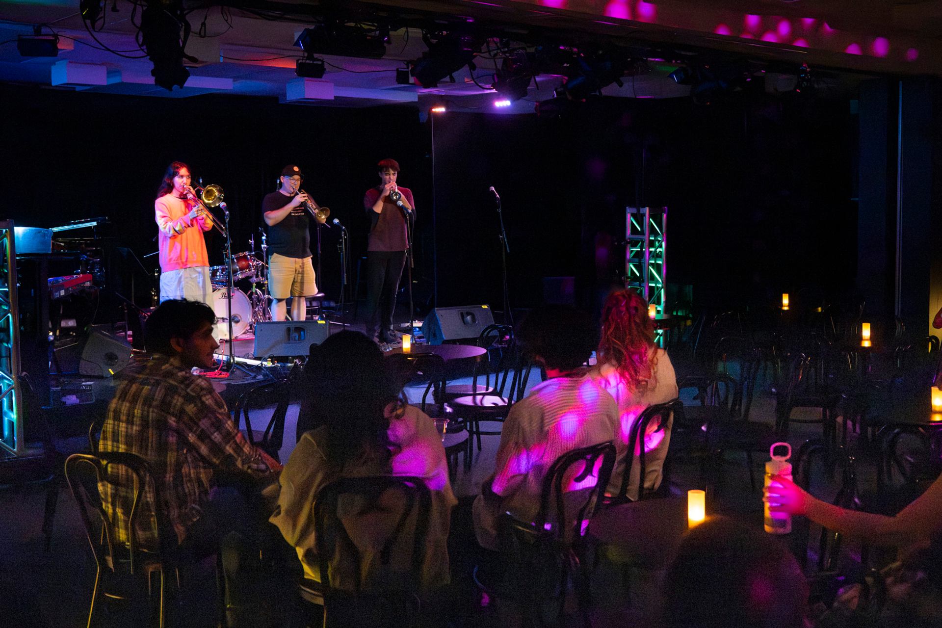 Small brass band performs on stage while seated audience watches under colourful lights.