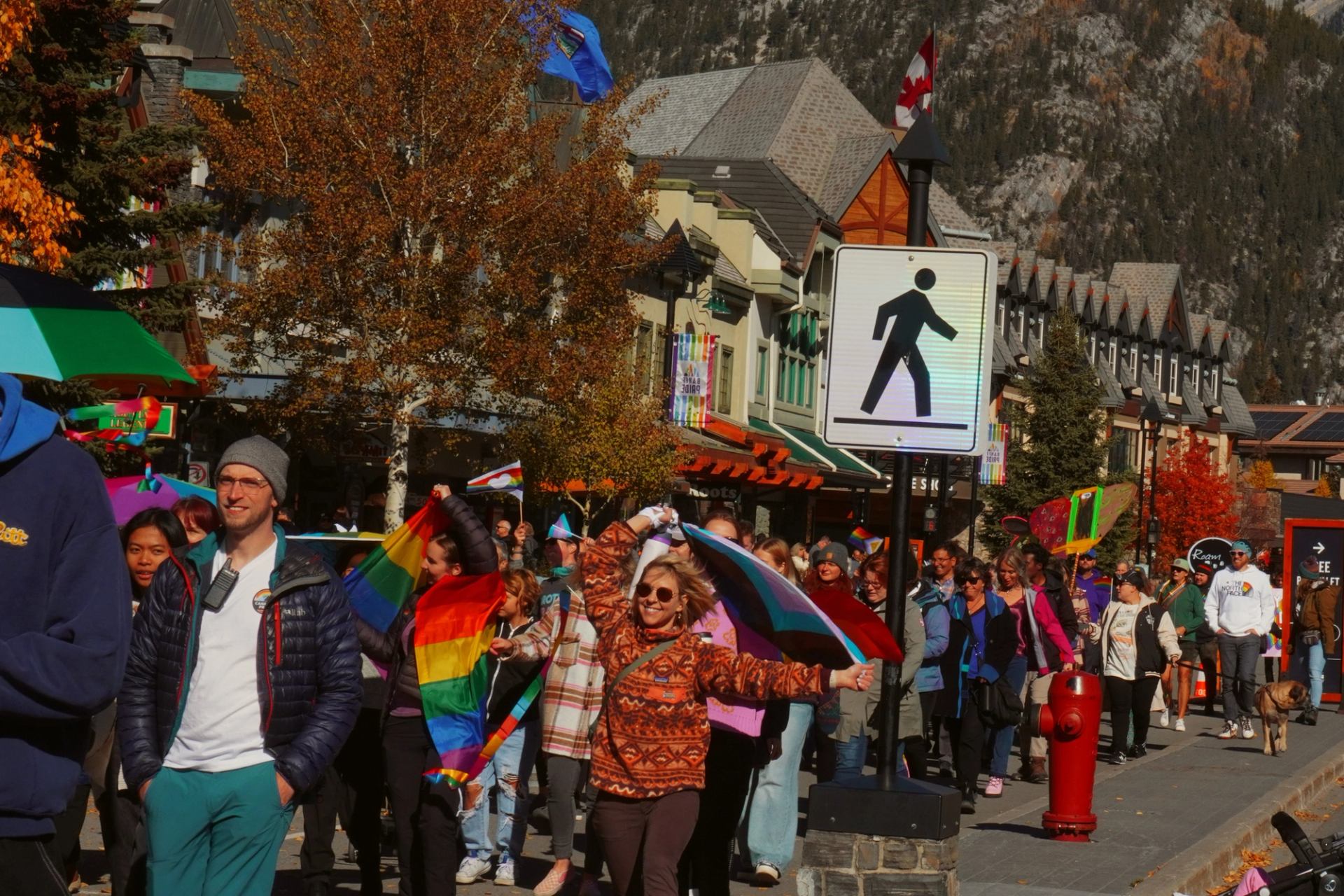 Banff Pride parade with rainbow flags and banner, set against mountains and autumn trees.