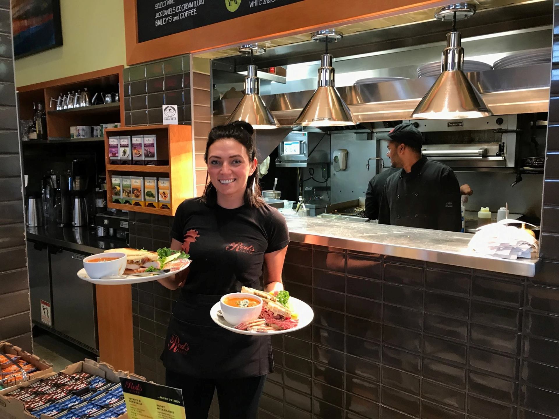 Smiling waitress holding two plates with food inside a diner kitchen.