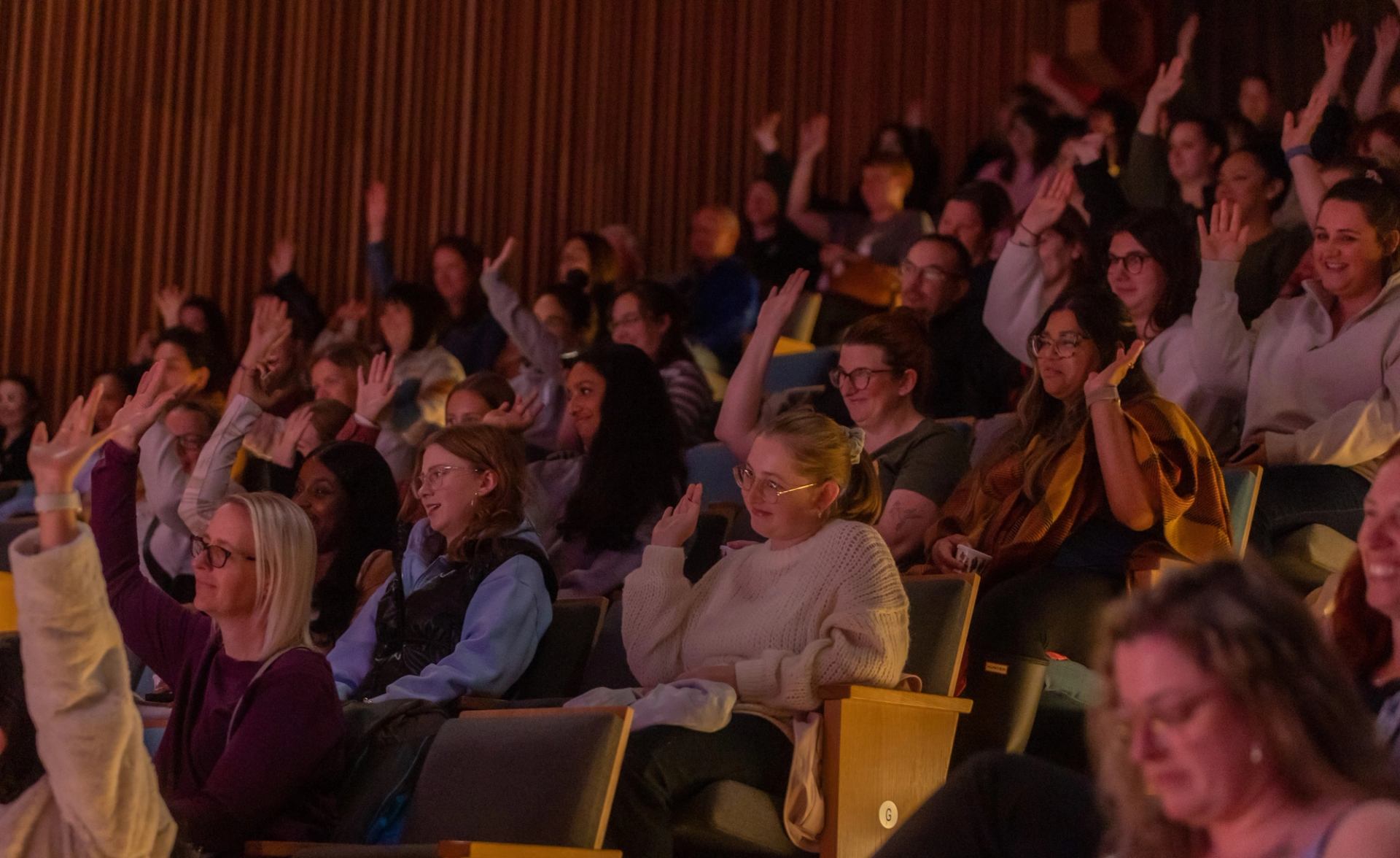 Theatre audience members raise their hands while seated in rows.