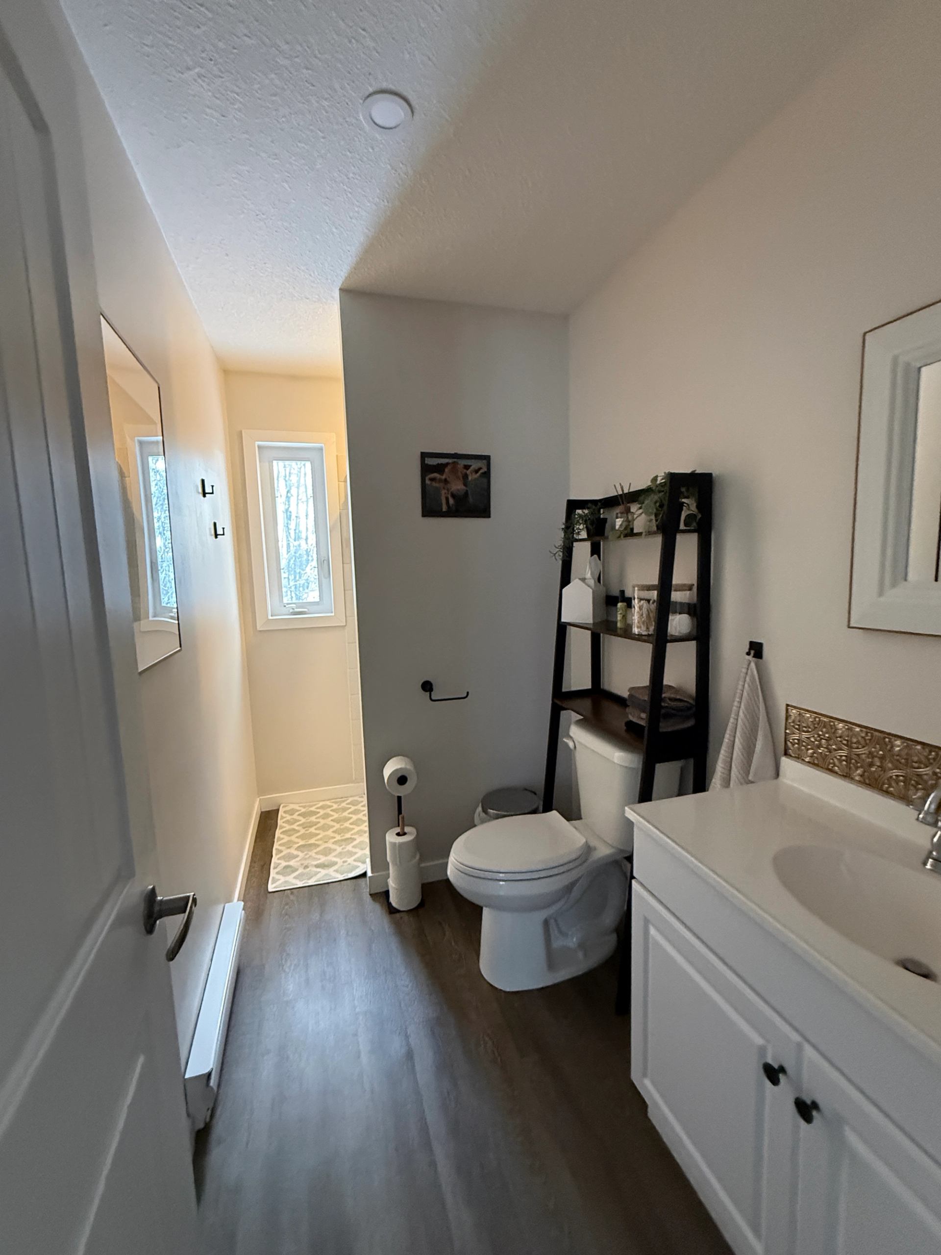 A clean bathroom with a white vanity, shelving unit, and a window letting in natural light.