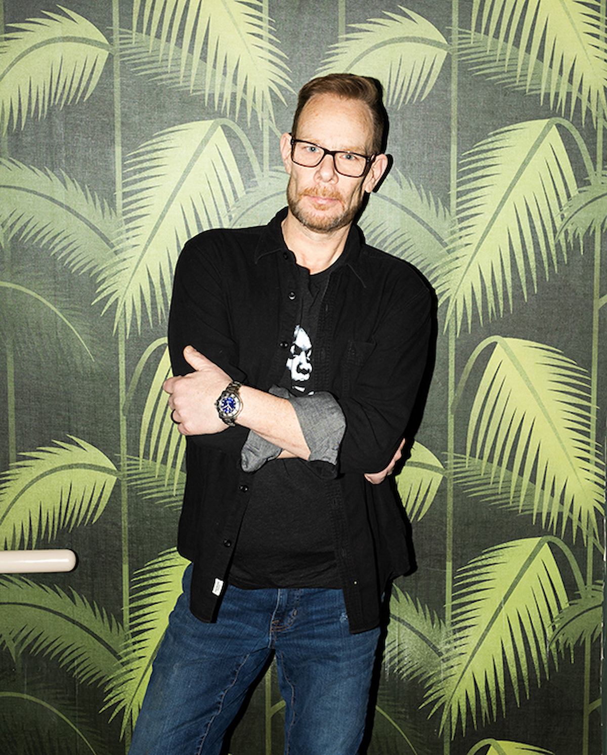 Man in glasses with arms crossed, standing in front of tropical leaf backdrop.