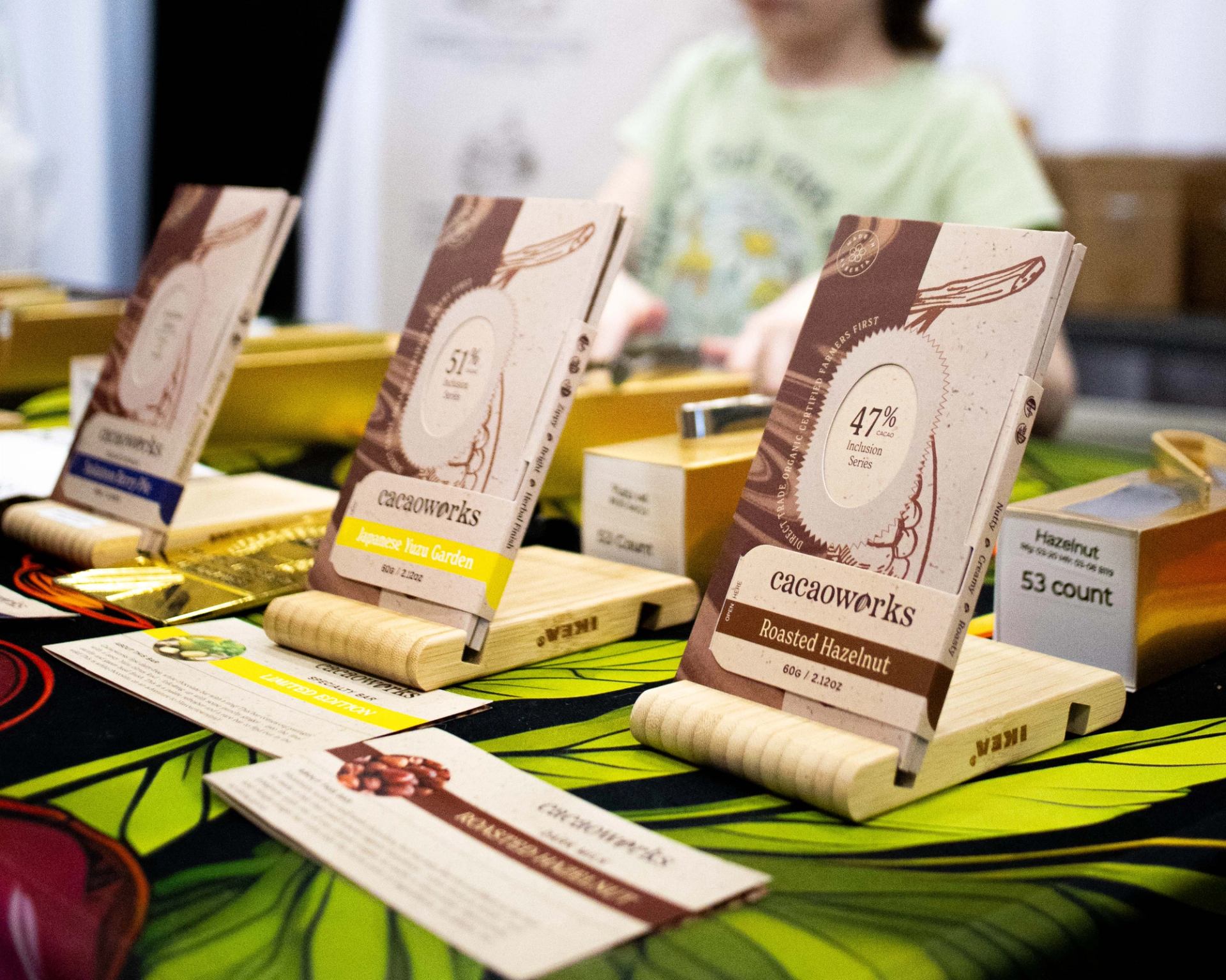 Display table with Cacaoworks chocolate bars on wooden stands and product information cards.