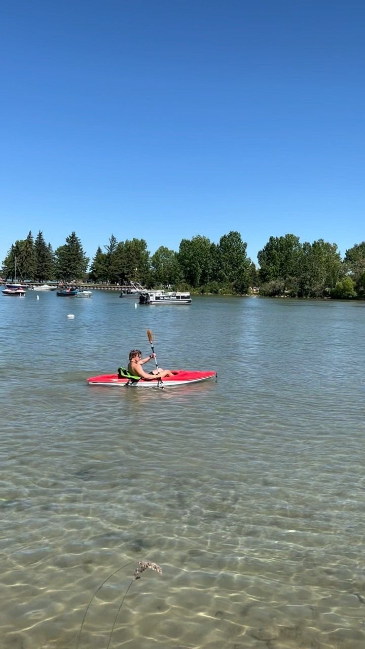 Person kayaking on clear water with trees and others enjoying water activities in background.
