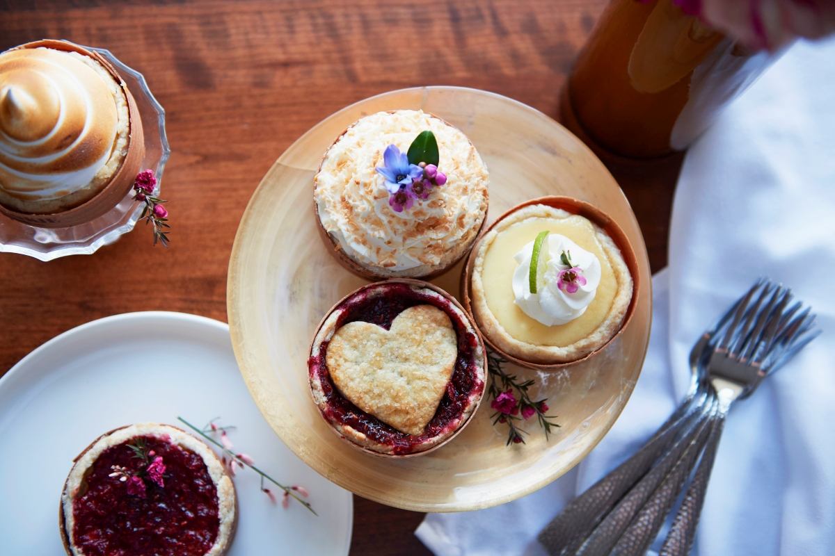 Top view of assorted mini pies decorated with fruit and flowers on wooden and ceramic plates.