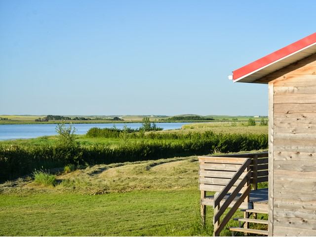 Wooden cabin deck overlooking grassy fields and a calm lake under a clear blue sky.
