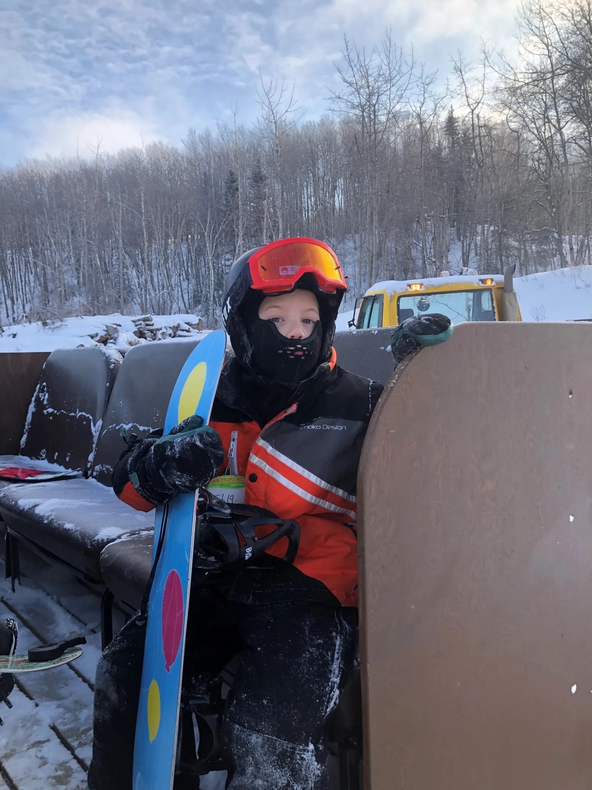 Snowboarder in orange jacket sits on snowy bench, holding board beside forested hill.