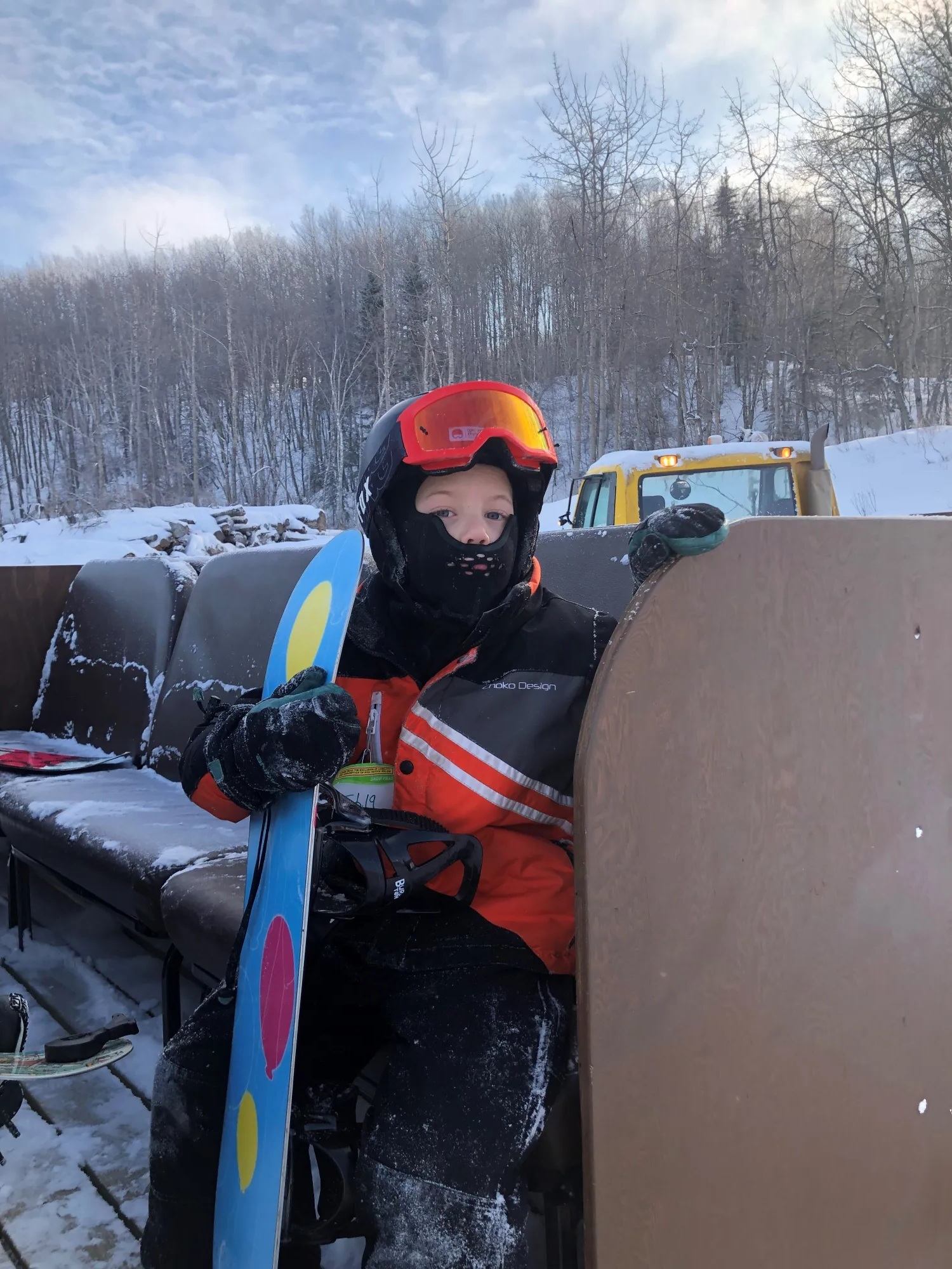 Snowboarder in orange jacket sits on snowy bench, holding board beside forested hill.