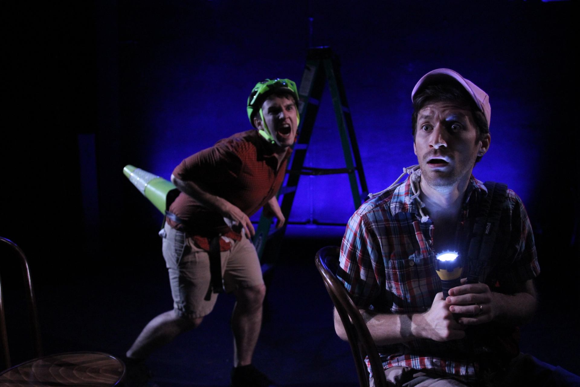 Stage scene with two actors, one holding flashlight, one wearing helmet and traffic cone.