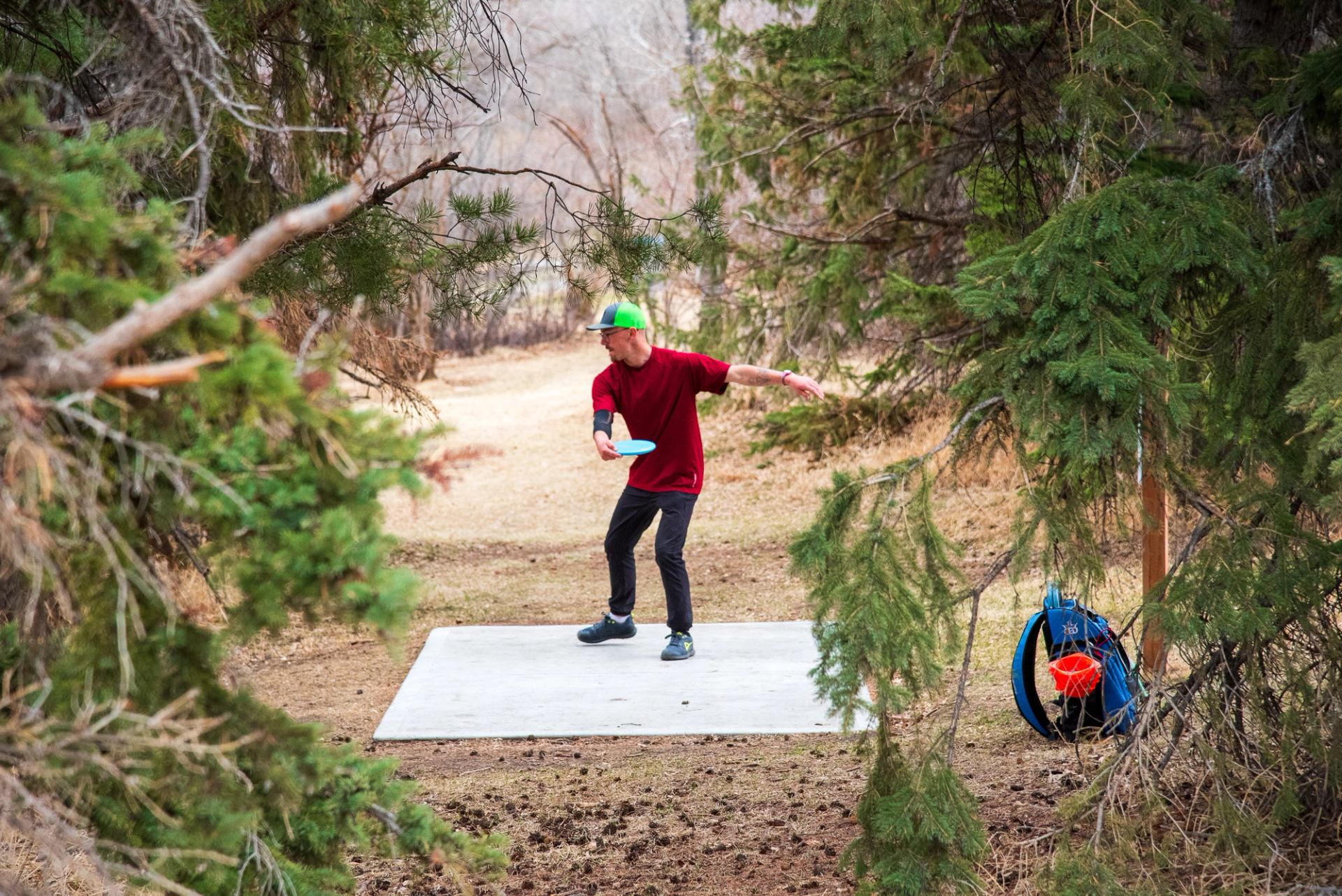 A young adult throwing a disc from the concrete tee pad of hole 6, a narrow fairway with lots of trees, on the Stoney Creek Disc Golf Course