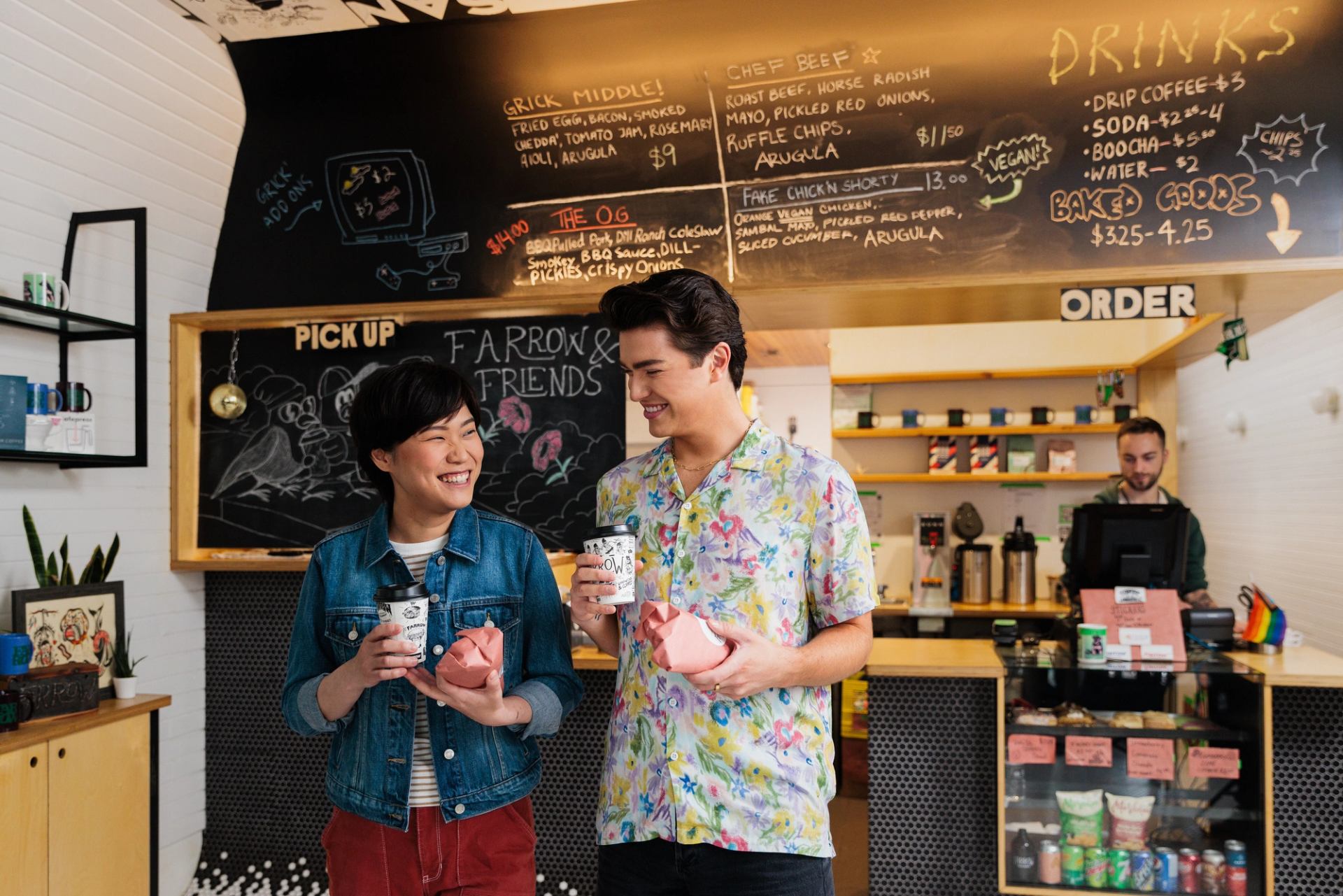 Café customers hold takeaway drinks and wrapped food beneath a chalkboard menu.