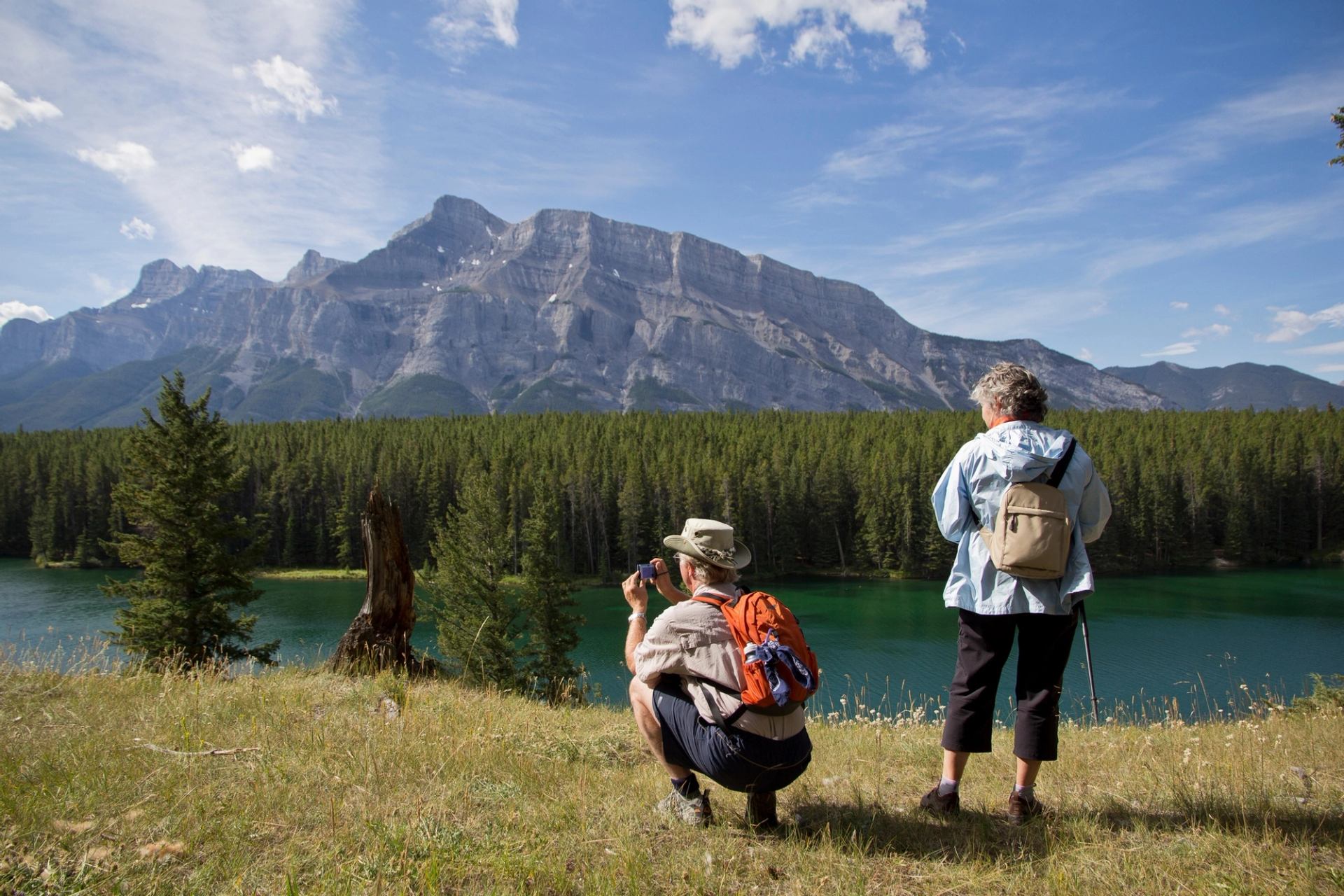 Two people overlook a turquoise lake, dense forest, and large mountains. One crouches to take a photo.