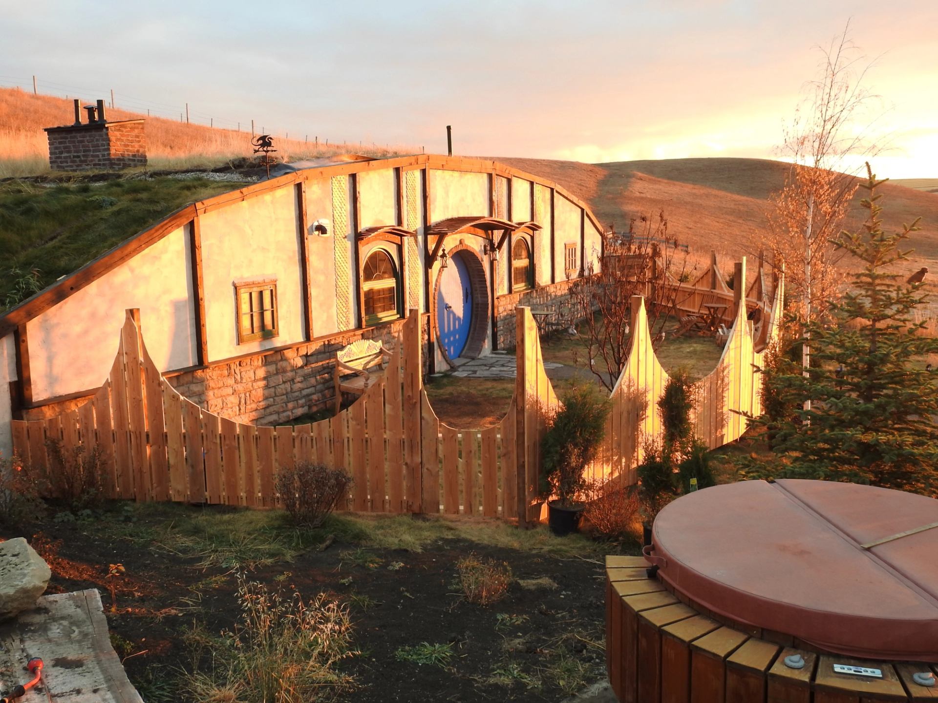 Charming hobbit-style home with round blue door, wood fence, and rolling hills at golden sunset.