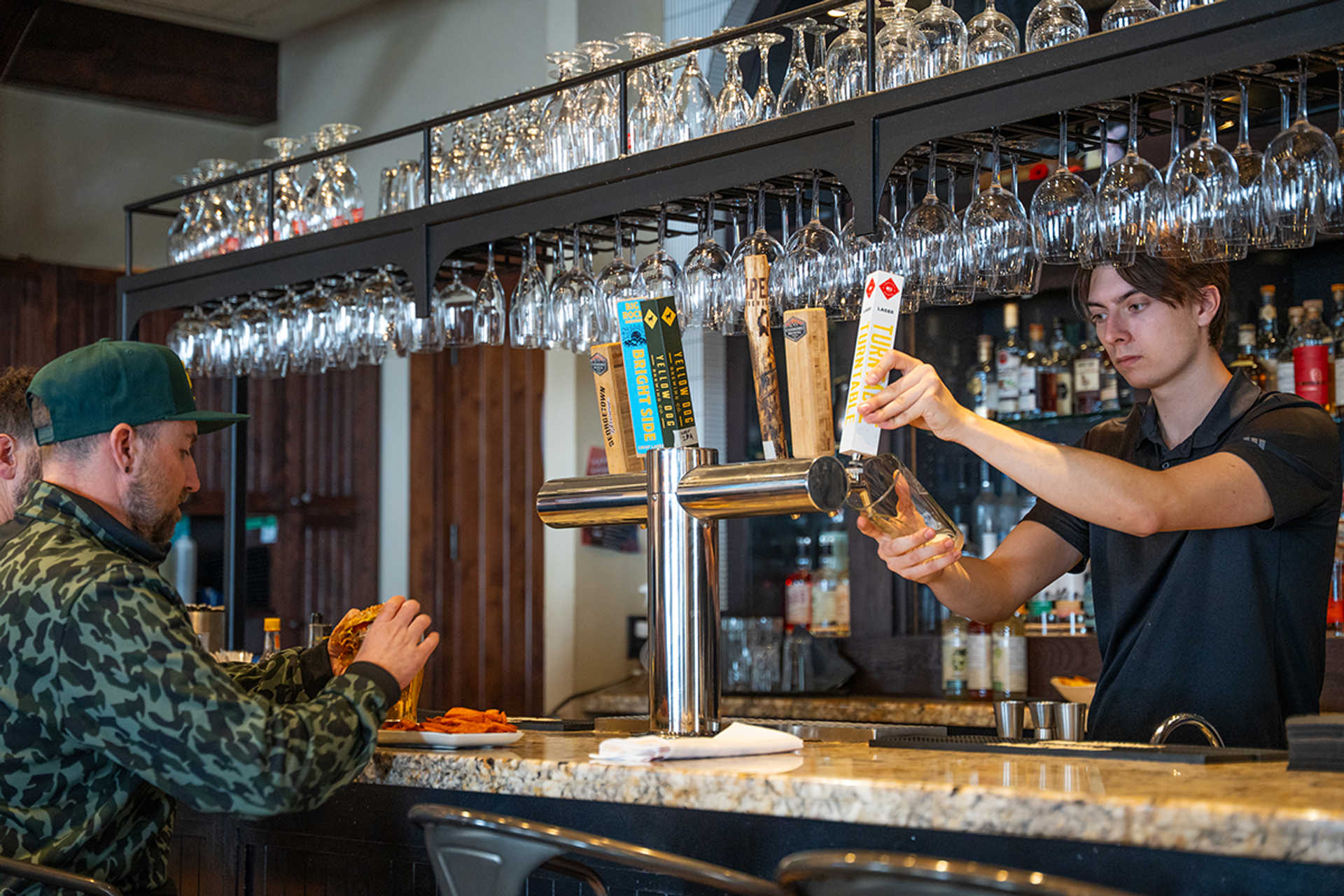 Bartender pours draft beer while customer eats at granite bar with hanging glasses.
