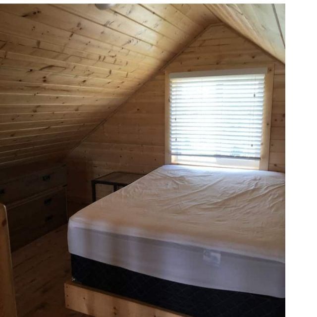 Cozy loft bedroom with wood-paneled walls, a sloped ceiling, and a bed near a window with blinds.